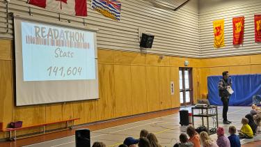 Elementary children sit on the floor of a school gymnasium looking at a screen that shows the school's read-a-thon statistics, and the school principal stands in front of that screen with a microphone.