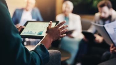 Person with tablet reviews financial charts while board members look on